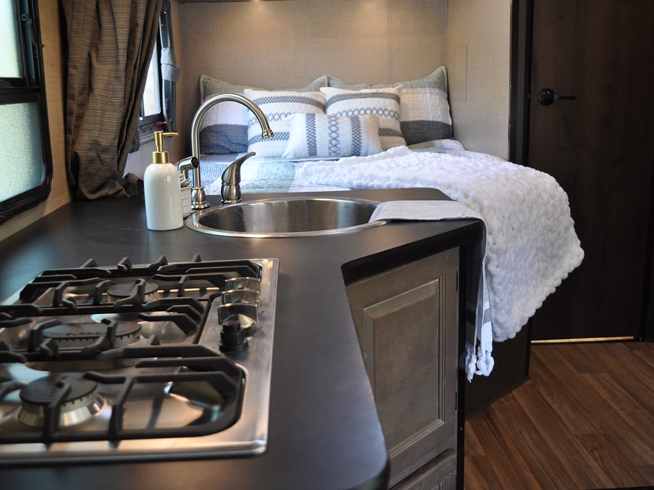 Kitchen area with open stove top, sink with towel, and assembled bed with pillows and quilt
